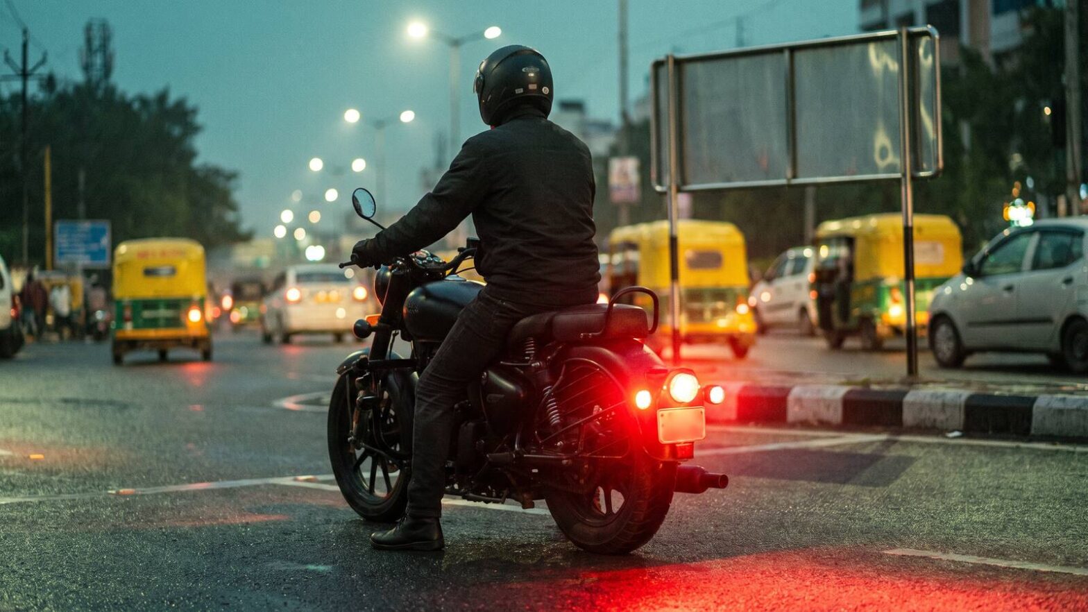 A motorcycle waiting at a wet Indian junction at dusk with a red auxiliary light glowing at the rear