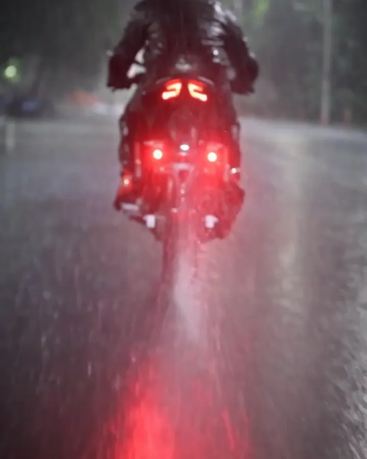Motorcycle riding away on a wet road at night in heavy rain with twin red strobe reflections trailing on the asphalt