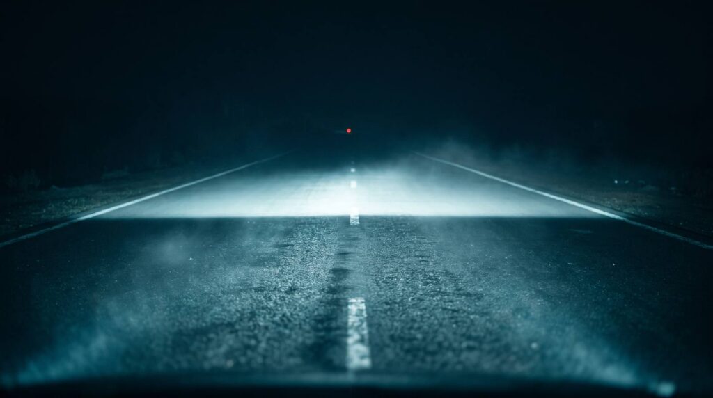 Night highway with a sharply cut-off white beam edge across wet tarmac and one distant red light ahead